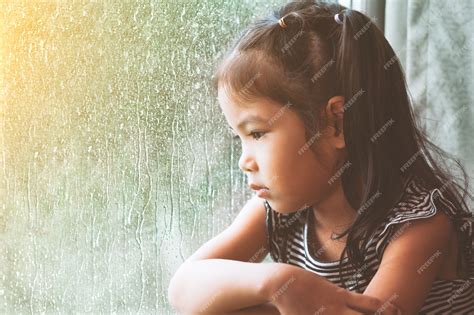 Premium Photo | Sad asian little girl looking outside through the window in the rainy day