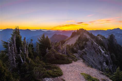 the sun is setting at the top of a mountain in the distance, with trees and rocks on the ground