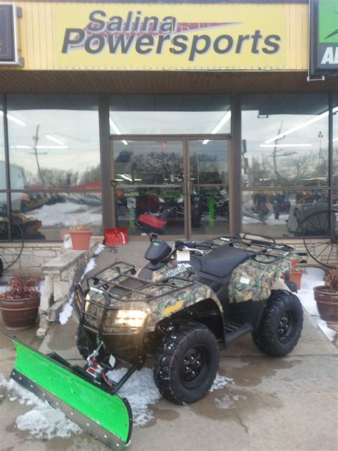 a green snow plow sitting in front of a power sports store on a snowy day