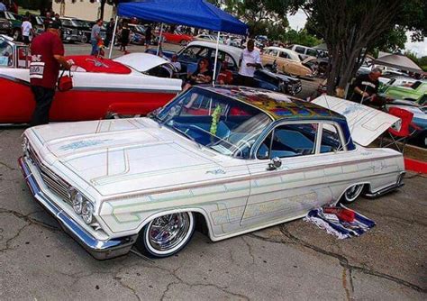 an old car is parked in a lot with other cars and people standing around it