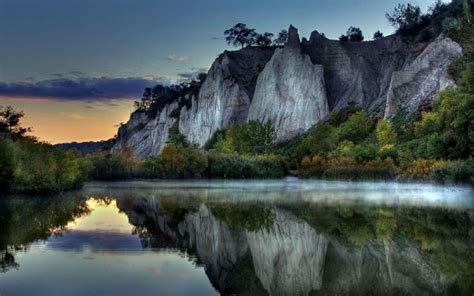 the sun is setting over some mountains and trees on the water's edge, with rocks in the background