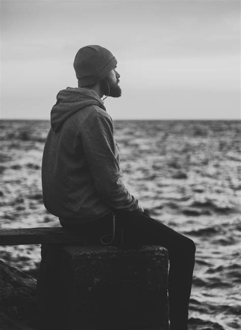Grayscale Photo of Man in Hoodie and Kit Cap Sitting Near Bodies of Water · Free Stock Photo