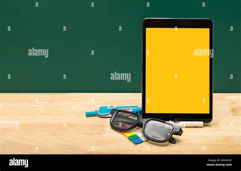 Blank yellow screen tablet with eyeglasses,pencil on wood table top with green blackboard ...