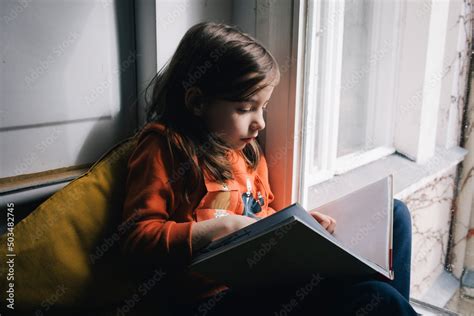 Girl reading book sitting by glass window at home Stock Photo | Adobe Stock