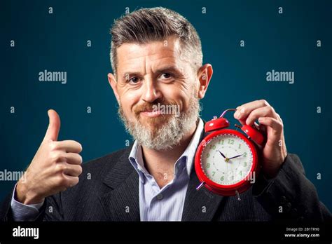 Bearded businessman in suit holding red alarm clock and showing thumb up gesture. Time ...