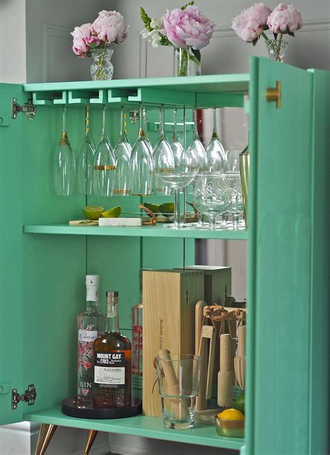a green cabinet with wine glasses and liquor bottles on it's shelf next to flowers