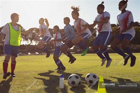 Side view of Caucasian coach helping diverse female soccer players with jumping exercise on the ...