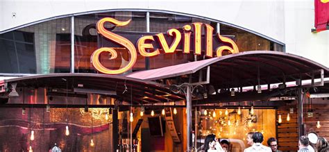 people are walking in front of a restaurant called sevillia, which is lit up at night