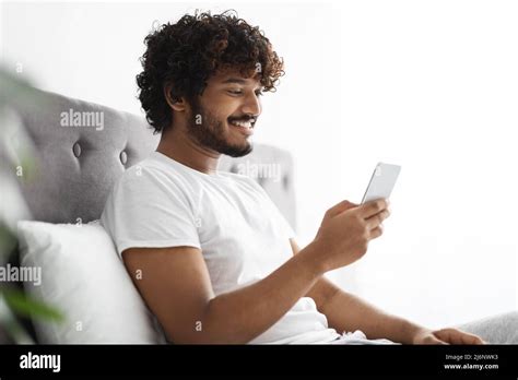 Positive indian guy scrolling in bed, using smartphone Stock Photo - Alamy