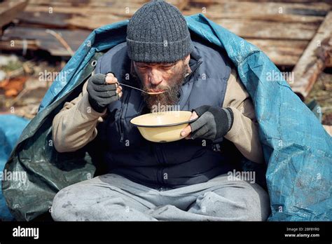 a homeless man eats soup from a plate near the ruins, helping poor and ...