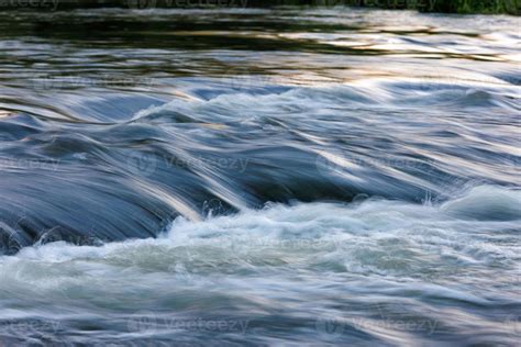 flowing water of a summer river with a small rapid waterfall at evening ...