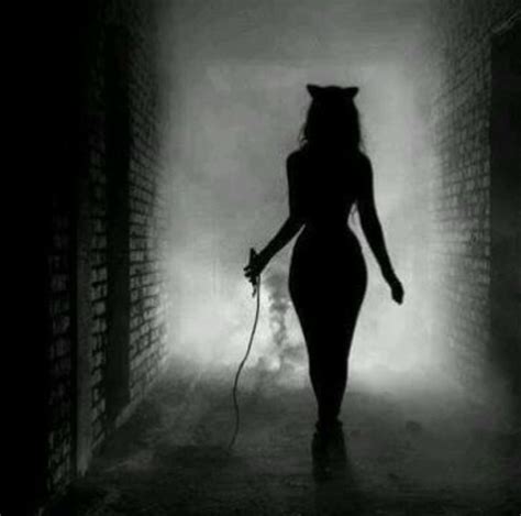 A Woman Walking Down A Dark Alley Way With A Cat On Her Head And A Whip