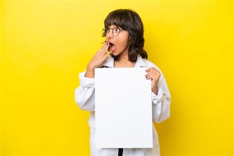 Jovem médica latina isolada em fundo amarelo segurando um cartaz vazio expressão de surpresa