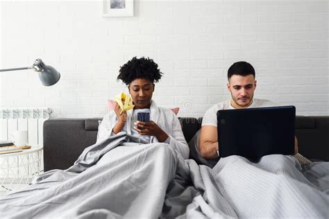 Young Multiracial Couple Using Their Mobile Electronic Devices In Bed