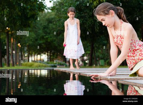 Girl Sitting By Edge Of Pond Touching Surface Of Water Another Girl In