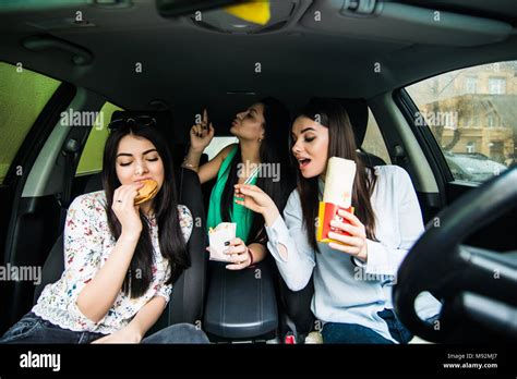 Three young women drive a car, speaking each other and eating a fast