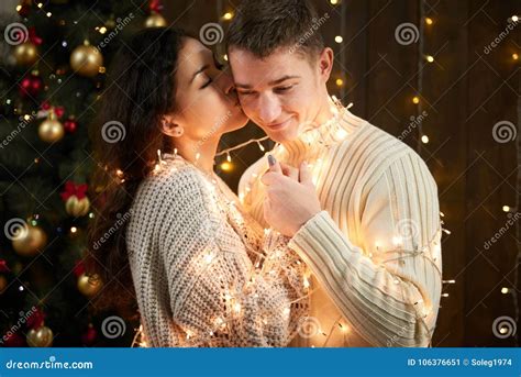 Young Couple Kissing In Christmas Lights And Decoration Dressed In White Fir Tree On Dark