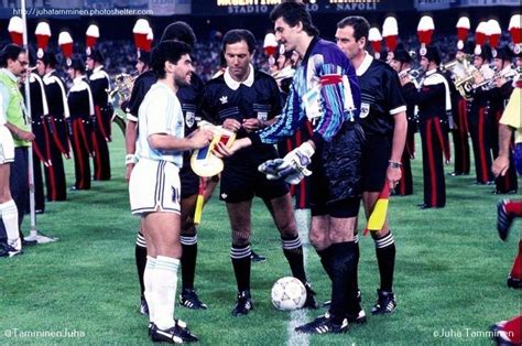 Two Men Shaking Hands Over A Soccer Ball On A Field With Other People