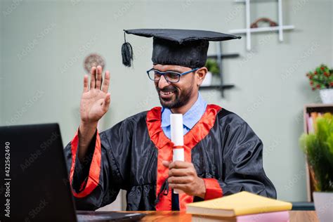 Student attending virtual graduation from laptop by holding certificate in graduation dress from ...