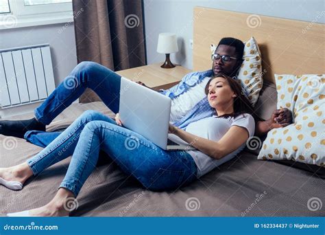 International Couple Smiling Looking At Laptop While Resting On Bed In