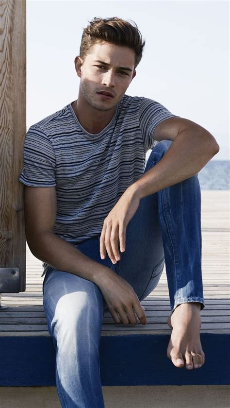 A Man Is Sitting On A Bench By The Water Wearing Jeans And A Striped T