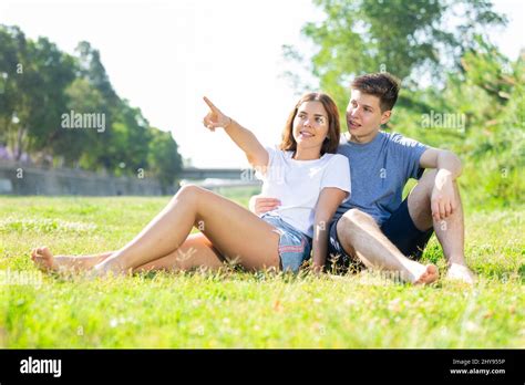 Happy Romantic Couple Sitting On Grass And Points To Something In Day