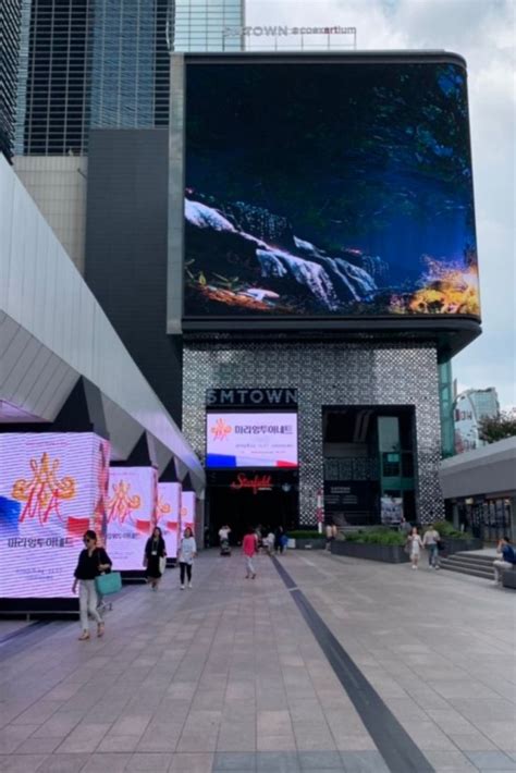 People Are Walking Around In Front Of A Building With A Large Screen On