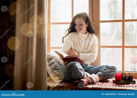 Brunette Woman Wearing Sweater And Festive Socks Reading Book Sitting On Windowsill In Room