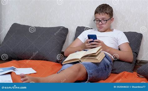 Teen In Glasses With A Smartphone In Hands And Open Book Studying