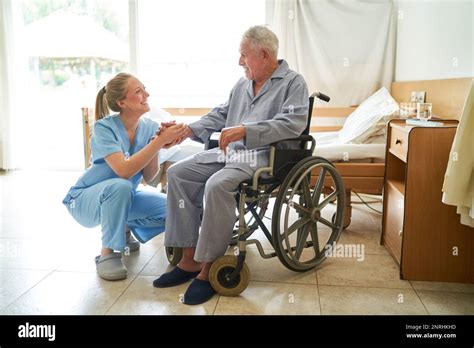 Smiling Caring Nurse Holding Hand Of Senior Man Sitting In Wheelchair At Rehab Center Stock