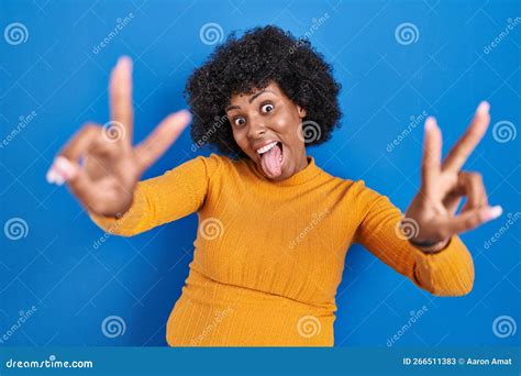 Black Woman With Curly Hair Standing Over Blue Background Smiling With Tongue Out Showing