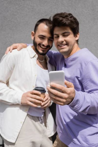 borrosa gay hombre abrazando sonriente novio en casual ropa con café a ir y el uso de móvil