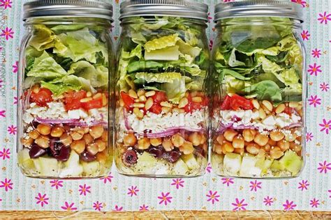 Three Mason Jars Filled With Salads On Top Of A Floral Table Cloth And Pink Flowers