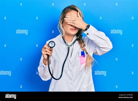 Beautiful Young Blonde Doctor Woman Holding Stethoscope Covering Eyes