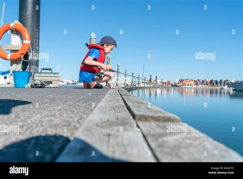 Boy sits on dock fishing wearing red life-jacket looking over edge into ...