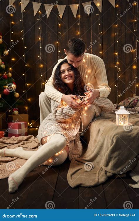 Young Couple Is In Christmas Lights And Decoration Dressed In White Fir Tree On Dark Wooden