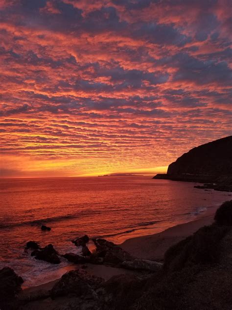 The Sun Is Setting Over The Ocean With Rocks And Cliffs In The Foreground