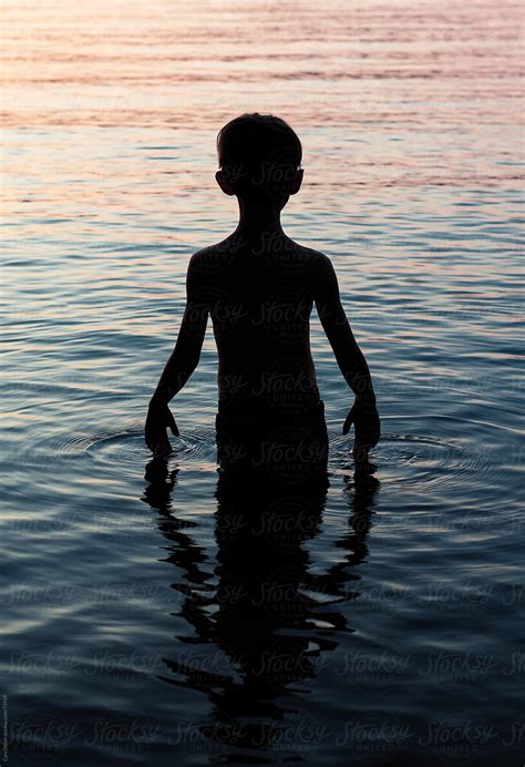 Silhouette Of A Boy Standing In Lake Water At Sunset By Stocksy