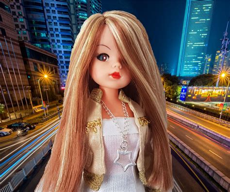 A Doll With Long Blonde Hair Standing In Front Of A Cityscape At Night