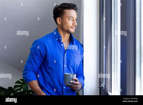 Happy Biracial Man Holding Mug And Looking Through Window Stock Photo