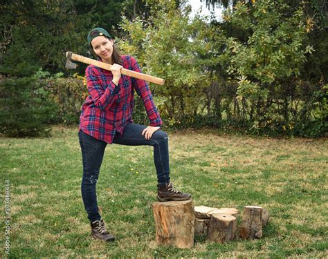 Smiling female lumberjack holding a huge axe on her shoulder standing with one leg on the log ...