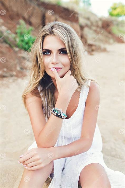 Attractive Blonde Girl With Long Hair And Blue Eyes Is Sitting On Beach She Wears White Dress