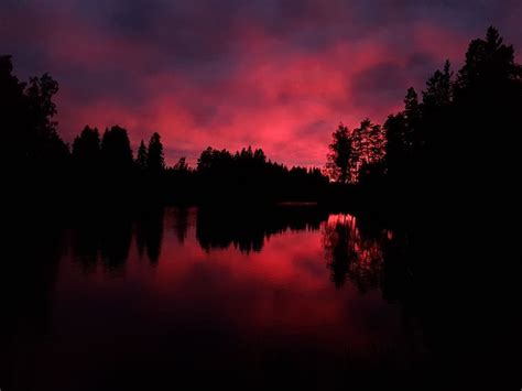 The Sun Is Setting Over A Lake With Trees In The Foreground And Clouds