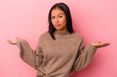 Joven mujer latina aislada sobre fondo rosa dudando y encogiéndose de