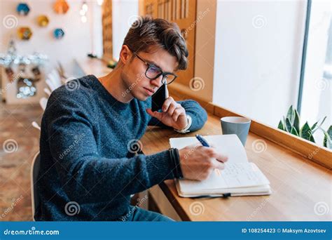 Man Writing Down Something In Notebook While Talking On Mobile Phone At