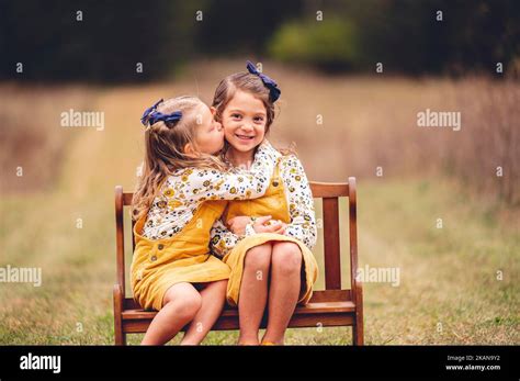 A Pair Of Young Twin Girls Dressed Identically In Yellow Kissing Each Other On The Cheek In A