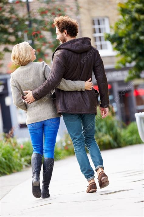 Rear View Of Young Couple Walking Through City Park Together Background