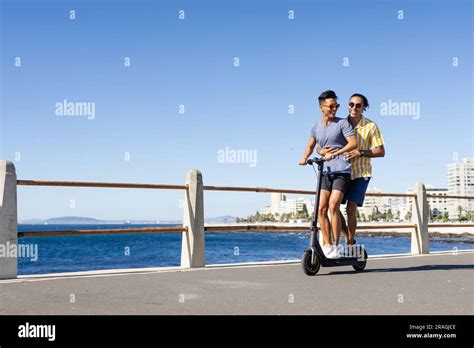 Happy Biracial Gay Male Couple Riding Scooter Together On Promenade By The Sea Copy Space Stock