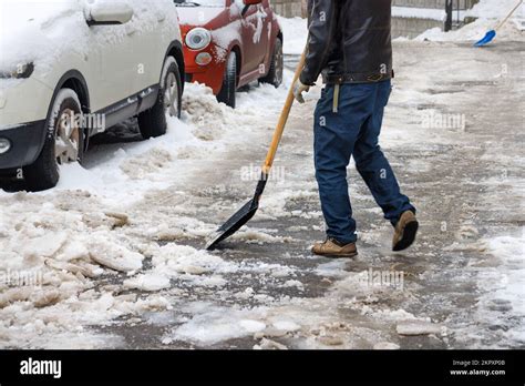 A Man Cleans Snow And Ice With A Shovel From An Asphalt Path Near