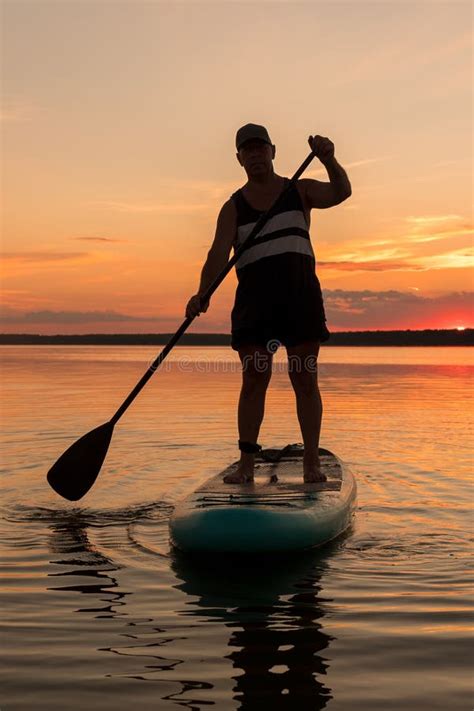 A Man In Shorts Stands On A Sup Board In The Evening At Sunset In The